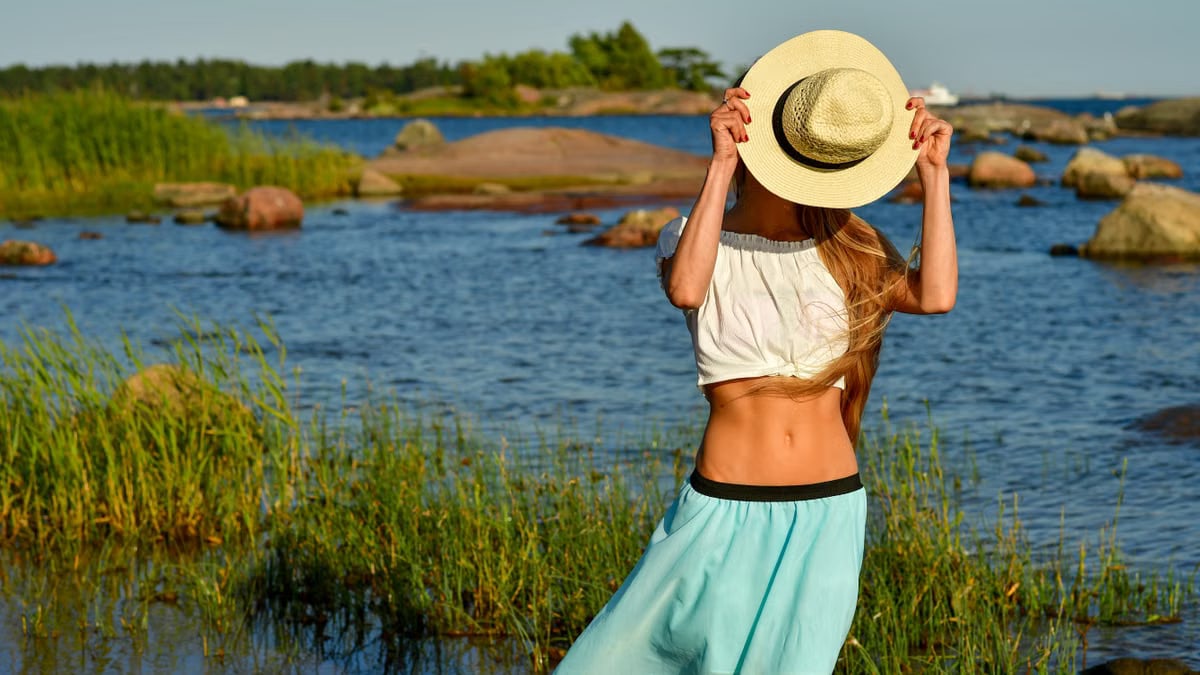 Woman enjoys a summer day by the rocky beach in Helsinki, hiding face with a straw hat